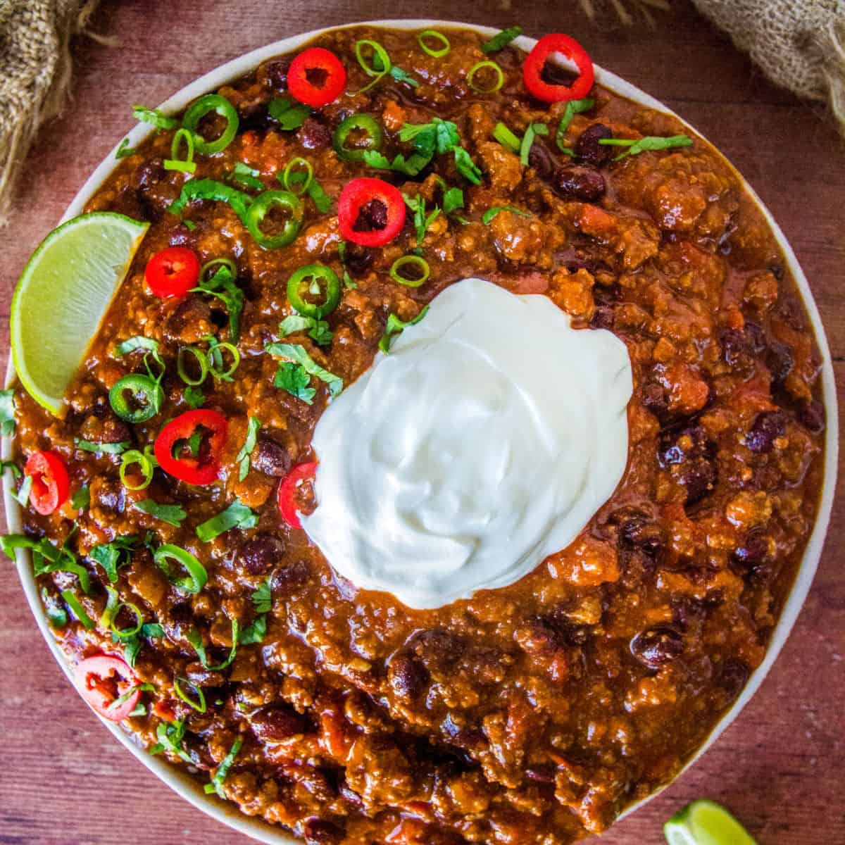 Bowl of Black Bean Chili Topped with Sour Cream, Red and Green Jalapeno Slices, Cilantro, and a Lime Wedge.