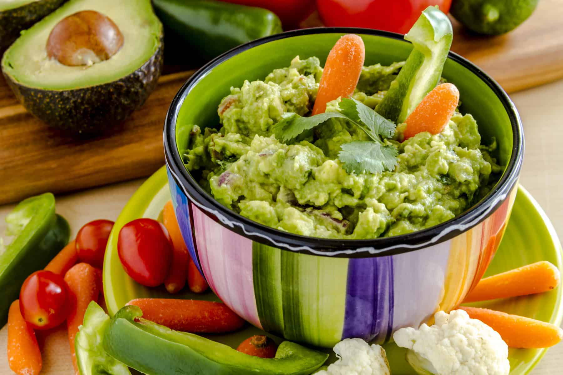 Fresh chunky guacamole in colorful bowl sitting on bright green plate garnished with raw carrots and green peppers and cilantro surrounded by raw vegetables
