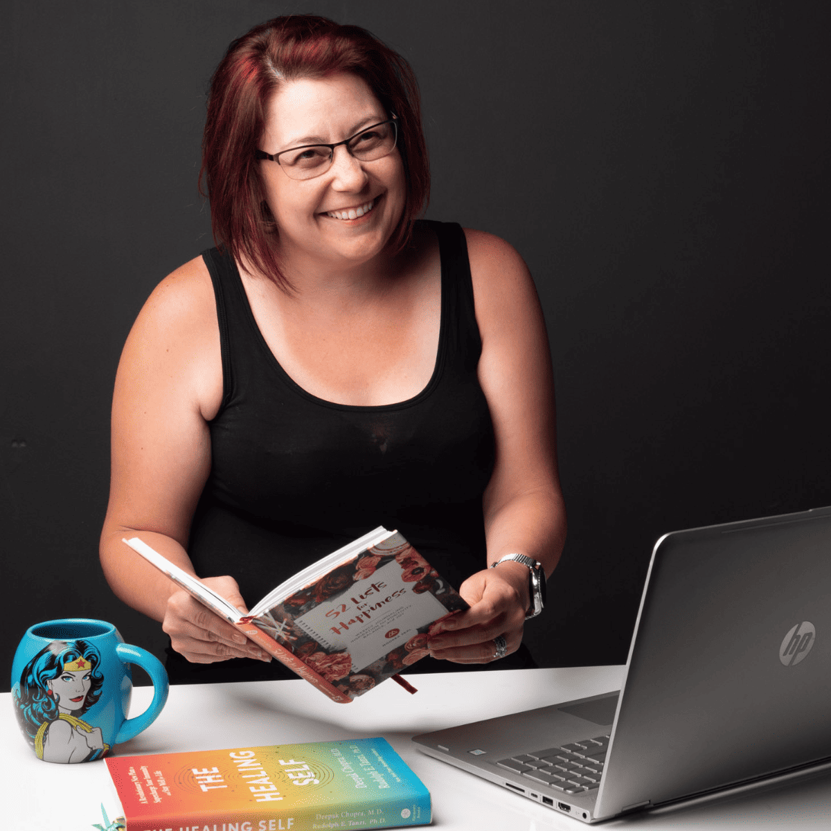 Heather Cooan reading a book and working on an HP laptop with a wonder woman mug and book on the table.