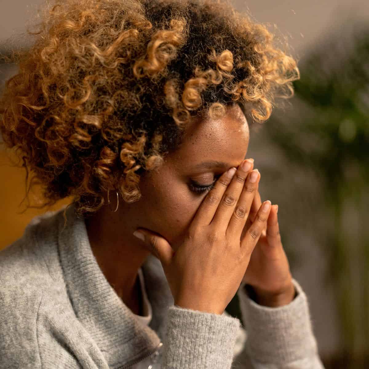 black woman wearing grey sweater sitting at a table with her head in her hands.