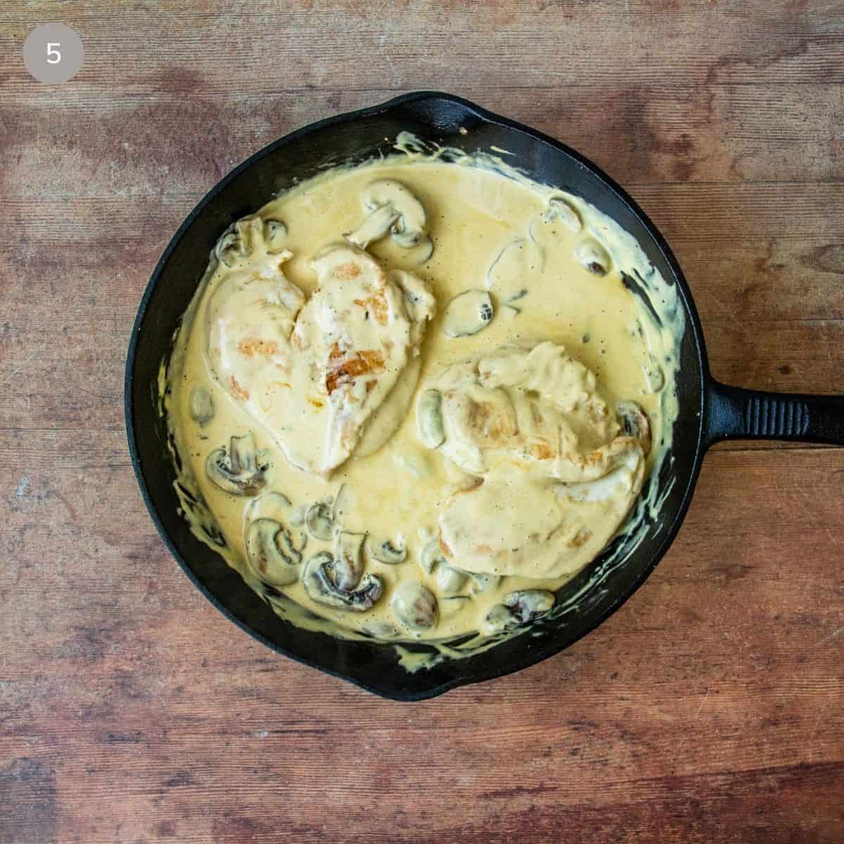 Chicken breasts simmering in creamy Dijon mushroom sauce in a cast iron skillet on wood.